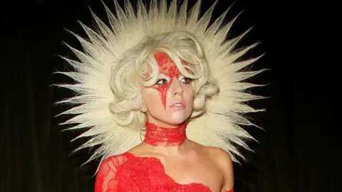 Getty Images Lady Gaga digambarkan pada tahun 2009. Dia mengenakan gaun renda merah dari bahu dengan renda yang juga dikenakan di lehernya dan di dahinya. Rambutnya melengkung dalam bob pirang yang diputihkan dan rambut yang terbuat dari rambut berbentuk seperti halo. 