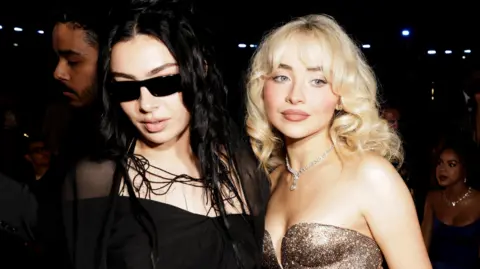 Getty Images Charli XCX dan Sabrina Carpenter yang digambarkan bersama di Grammy's. Charli memiliki rambut hitam panjang yang longgar dan pasangan kacamata hitam gelap dengan gaun bertali hitam. Sabrina memiliki rambut pirang di ikal khasnya dan mengenakan gaun emas tanpa tali. 